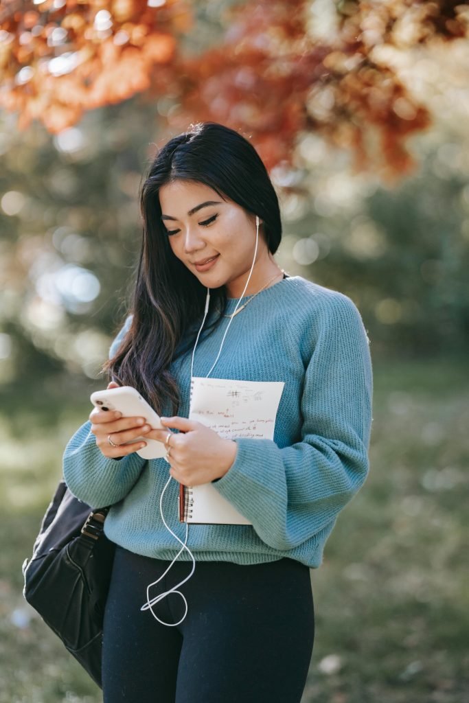 pexels-photo-5965925-5965925 Smiling woman with earbuds using smartphone and notebook in a sunny park.